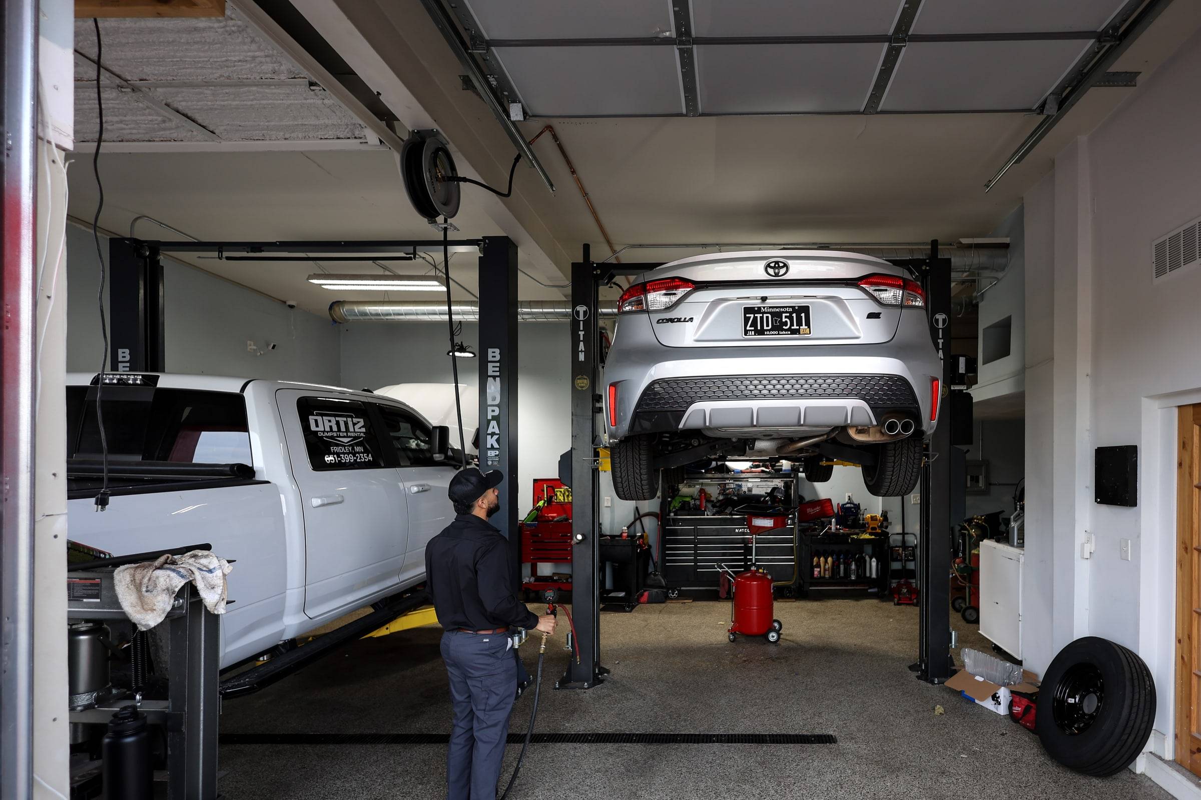 A car raised on the lift in one of the two bays, with the lead tech standing beside it and a pickup in the other bay