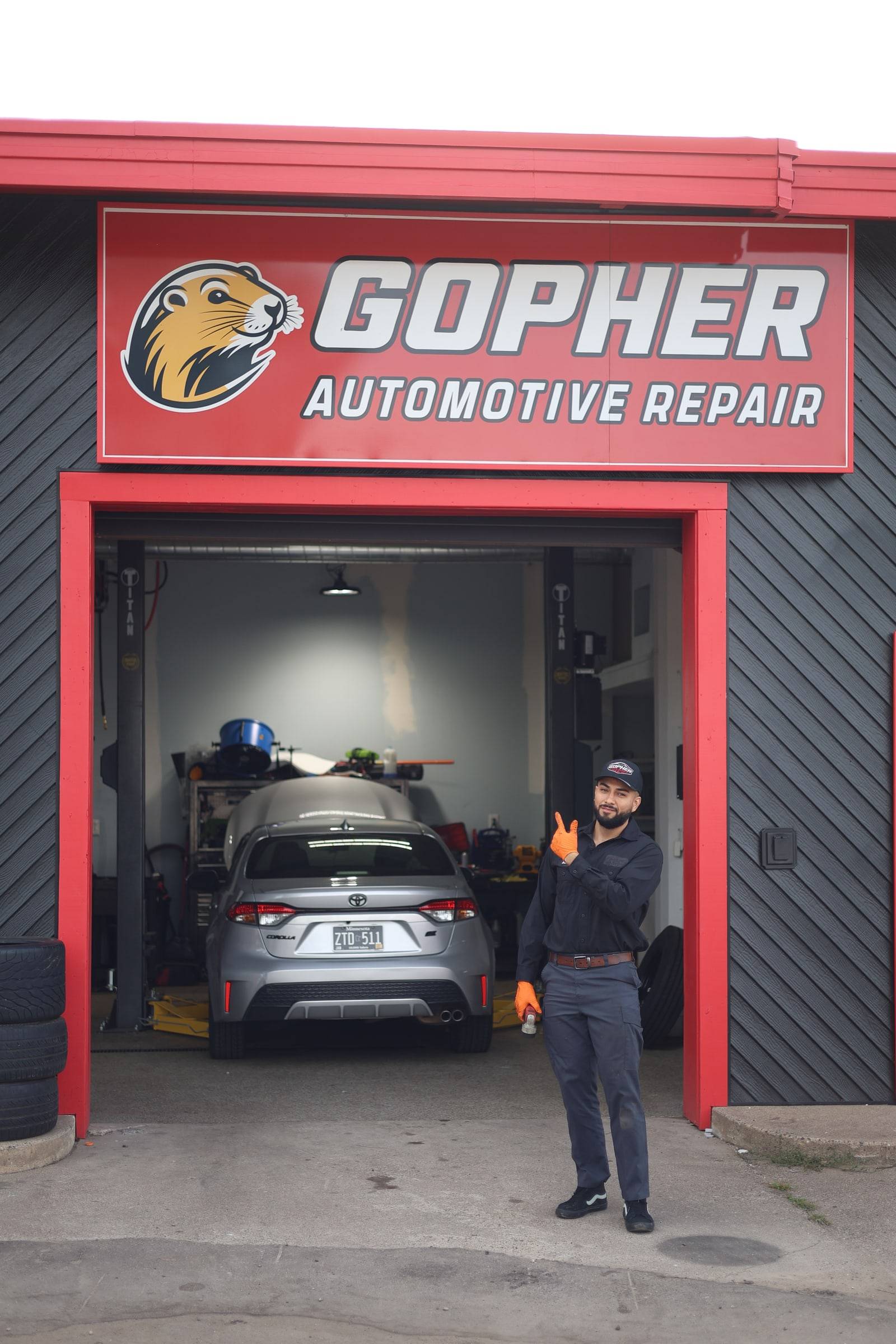 The lead tech standing at an open red bay door under the Gopher sign, gesturing toward the car inside